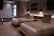 A modern hotel room features two neatly made double beds with striped pillows and runners. The room has a minimalist design with neutral tones, decorated with framed pictures and wall-mounted lights above the beds. A small seating area by a large window adds to the comfortable atmosphere.