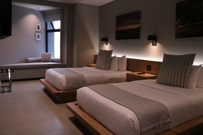 A modern hotel room features two neatly made double beds with striped pillows and runners. The room has a minimalist design with neutral tones, decorated with framed pictures and wall-mounted lights above the beds. A small seating area by a large window adds to the comfortable atmosphere.