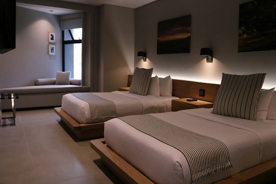 A modern hotel room features two neatly made double beds with striped pillows and runners. The room has a minimalist design with neutral tones, decorated with framed pictures and wall-mounted lights above the beds. A small seating area by a large window adds to the comfortable atmosphere.