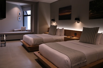 A modern hotel room features two neatly made double beds with striped pillows and runners. The room has a minimalist design with neutral tones, decorated with framed pictures and wall-mounted lights above the beds. A small seating area by a large window adds to the comfortable atmosphere.