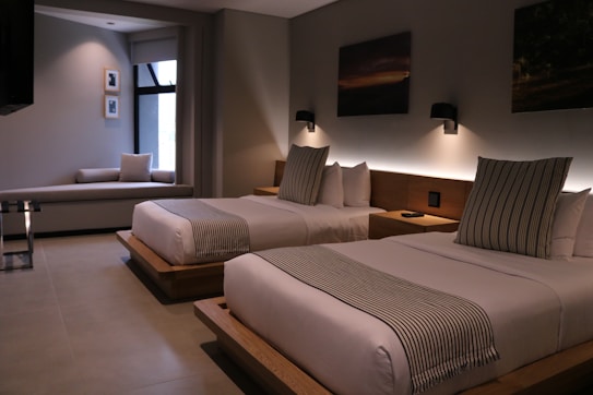 A modern hotel room features two neatly made double beds with striped pillows and runners. The room has a minimalist design with neutral tones, decorated with framed pictures and wall-mounted lights above the beds. A small seating area by a large window adds to the comfortable atmosphere.