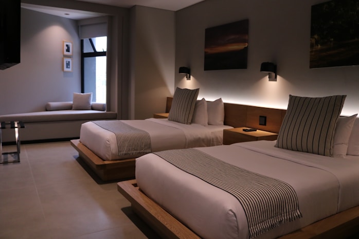 A modern hotel room features two neatly made double beds with striped pillows and runners. The room has a minimalist design with neutral tones, decorated with framed pictures and wall-mounted lights above the beds. A small seating area by a large window adds to the comfortable atmosphere.