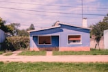 A small blue house with a gabled roof is situated at the center of a grassy yard. The front of the house has a window with closed shutters and a door. The walls are blue with a red brick base. There are trees and bushes surrounding the property, with a clear blue sky above. Power lines run across the top portion of the image.