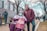 A person in a wheelchair and another standing person, both dressed warmly, are participating in a public outdoor event. The person in the wheelchair is holding a sign that reads 'Consistently Pro-Life' and is wearing a face mask and winter hat. The standing person is wearing glasses and a maroon jacket. Other people and signs are visible in the background, along with a paved walkway and bare trees, indicating it's likely winter.