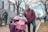 A person in a wheelchair and another standing person, both dressed warmly, are participating in a public outdoor event. The person in the wheelchair is holding a sign that reads 'Consistently Pro-Life' and is wearing a face mask and winter hat. The standing person is wearing glasses and a maroon jacket. Other people and signs are visible in the background, along with a paved walkway and bare trees, indicating it's likely winter.