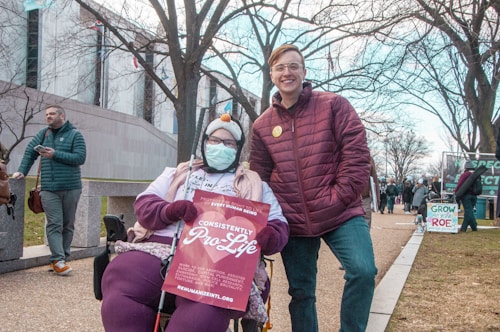 A person in a wheelchair and another standing person, both dressed warmly, are participating in a public outdoor event. The person in the wheelchair is holding a sign that reads 'Consistently Pro-Life' and is wearing a face mask and winter hat. The standing person is wearing glasses and a maroon jacket. Other people and signs are visible in the background, along with a paved walkway and bare trees, indicating it's likely winter.