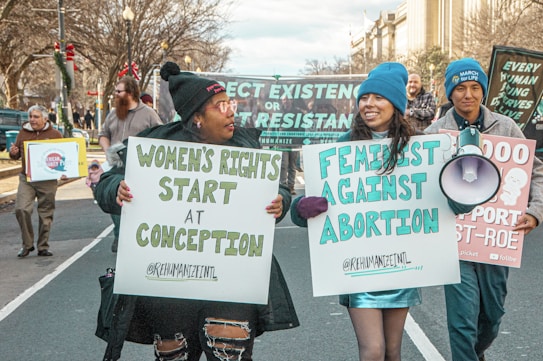 A group of people walking along a street holding signs related to anti-abortion messages. Prominent signs include statements like 'Women's Rights Start at Conception' and 'Feminist Against Abortion'. The participants are dressed warmly and appear to be in a peaceful protest or march setting. Buildings and trees are visible in the background.