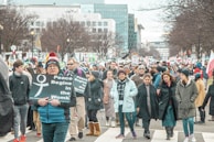 A large assembly of people is marching down a city street. Some participants are holding signs with messages, including 'Peace Begins in the Womb.' The group is diverse, composed of men and women of various ages, dressed in winter clothing. The backdrop features urban buildings and bare trees, indicating a colder season.