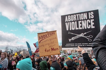 A large group of people, many holding signs, participate in a public demonstration. The signs have anti-abortion messages, including one that reads 'ABORTION IS VIOLENCE' and another that mentions abortion as an easy way out for men. The crowd is diverse, with people dressed in winter clothing, suggesting cold weather conditions.