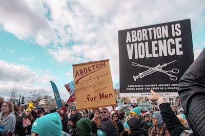 A large group of people, many holding signs, participate in a public demonstration. The signs have anti-abortion messages, including one that reads 'ABORTION IS VIOLENCE' and another that mentions abortion as an easy way out for men. The crowd is diverse, with people dressed in winter clothing, suggesting cold weather conditions.