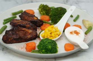 A plate with grilled chicken pieces accompanied by steamed broccoli, carrot slices, and green beans. Corn kernels are served in a small white spoon, alongside a dollop of white creamy sauce sprinkled with paprika. The arrangement suggests a nutritious, balanced meal.