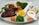 A plate with grilled chicken pieces accompanied by steamed broccoli, carrot slices, and green beans. Corn kernels are served in a small white spoon, alongside a dollop of white creamy sauce sprinkled with paprika. The arrangement suggests a nutritious, balanced meal.