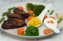 A plate with grilled chicken pieces accompanied by steamed broccoli, carrot slices, and green beans. Corn kernels are served in a small white spoon, alongside a dollop of white creamy sauce sprinkled with paprika. The arrangement suggests a nutritious, balanced meal.