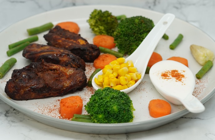 A plate with grilled chicken pieces accompanied by steamed broccoli, carrot slices, and green beans. Corn kernels are served in a small white spoon, alongside a dollop of white creamy sauce sprinkled with paprika. The arrangement suggests a nutritious, balanced meal.