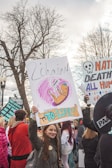 A lively scene of a protest or rally with several people holding signs. A prominent sign displays an illustration of a fetus with the words 'To Life!' and 'L'chaim'. The participants are bundled in winter clothing, suggesting a cold day. Bare trees and overcast skies add to the backdrop, while diverse signs express messages related to life and possibly anti-abortion sentiments.