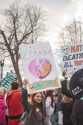 A lively scene of a protest or rally with several people holding signs. A prominent sign displays an illustration of a fetus with the words 'To Life!' and 'L'chaim'. The participants are bundled in winter clothing, suggesting a cold day. Bare trees and overcast skies add to the backdrop, while diverse signs express messages related to life and possibly anti-abortion sentiments.