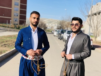 Two men stand on a sunny street with a backdrop of parked cars and buildings. Both are wearing traditional clothing; one is dressed in a royal blue outfit with a floral patterned belt, and the other in a gray outfit with dark sunglasses. The scene is set in an urban area with bare winter trees and distant hills.