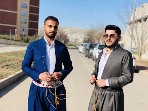 Two men stand on a sunny street with a backdrop of parked cars and buildings. Both are wearing traditional clothing; one is dressed in a royal blue outfit with a floral patterned belt, and the other in a gray outfit with dark sunglasses. The scene is set in an urban area with bare winter trees and distant hills.
