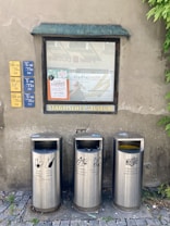 Metal recycling bins for different types of waste are placed in front of a wall with a window displaying posters. The wall also features signage with schedules and a label for the municipal museum. Lush green plants are partially visible on the right side.
