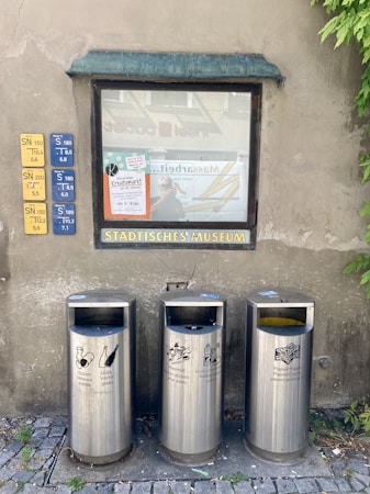 Metal recycling bins for different types of waste are placed in front of a wall with a window displaying posters. The wall also features signage with schedules and a label for the municipal museum. Lush green plants are partially visible on the right side.