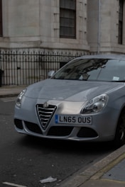 A silver Alfa Romeo car is parked along a city street next to a beige building with tall windows and a black iron fence. The vehicle's front license plate displays LN65 OUS. The street and building architecture suggest an urban setting.