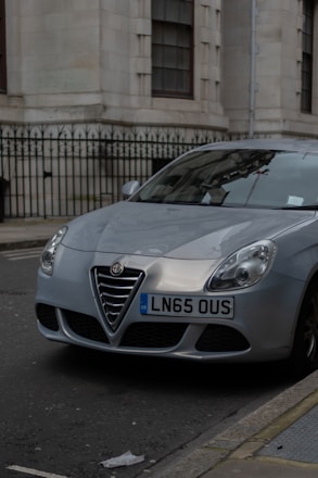 A silver Alfa Romeo car is parked along a city street next to a beige building with tall windows and a black iron fence. The vehicle's front license plate displays LN65 OUS. The street and building architecture suggest an urban setting.