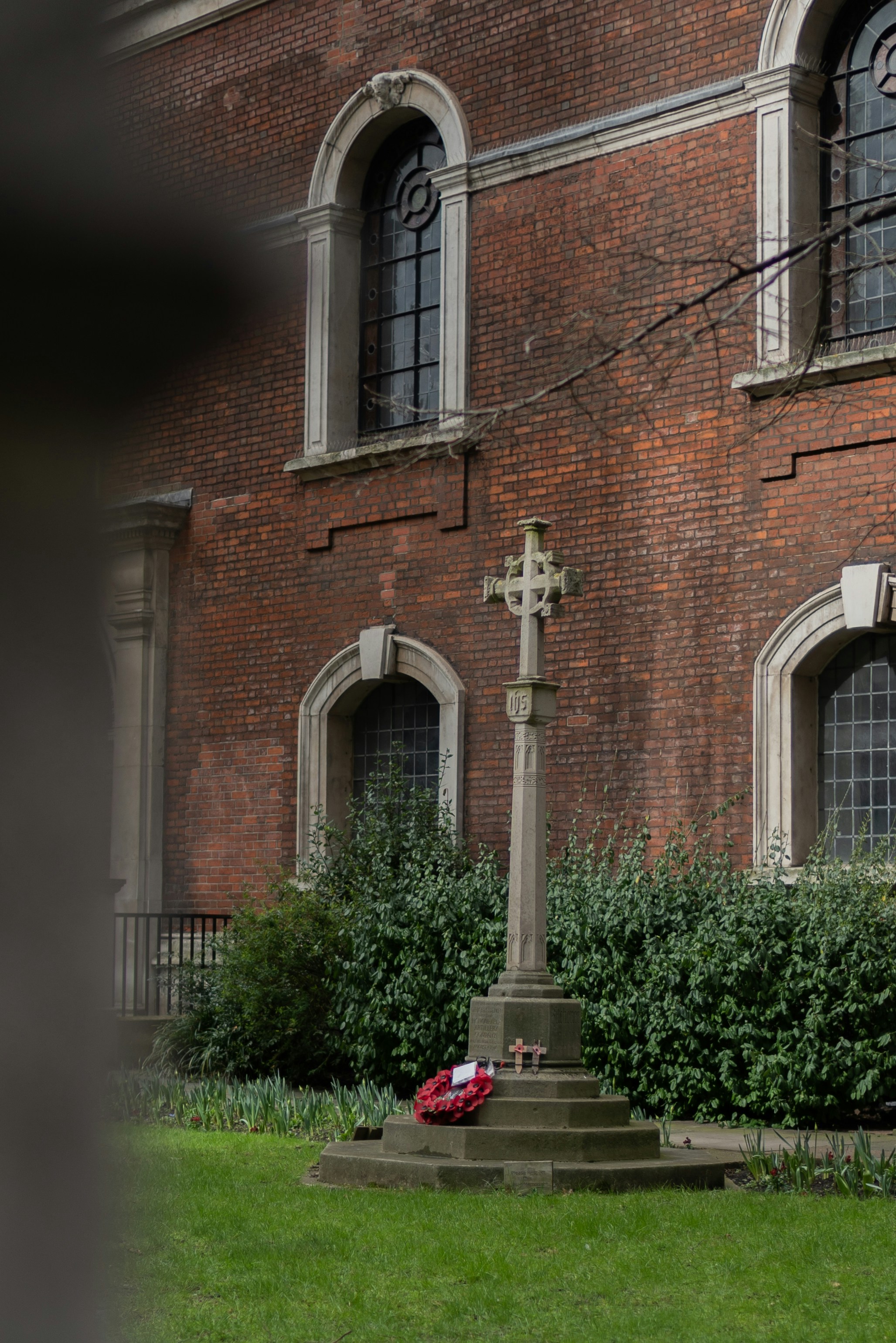 A stone cross monument on a pedestal with a wreath of red flowers placed at its base, situated in front of a red-brick building with arched windows. The surrounding area includes green grass and shrubs, enhancing the solemn atmosphere of the scene.