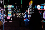 Dimly lit arcade scene with various brightly colored neon lights from different game machines. Several arcade machines, including Monopoly-themed and a large Connect Four game, line the room. The floor has a colorful carpet pattern, and overhead lights create a lively atmosphere.