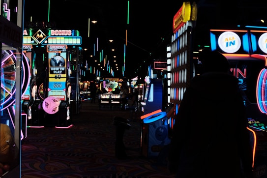 Dimly lit arcade scene with various brightly colored neon lights from different game machines. Several arcade machines, including Monopoly-themed and a large Connect Four game, line the room. The floor has a colorful carpet pattern, and overhead lights create a lively atmosphere.