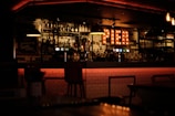 A warmly lit bar interior with shelves stocked with various bottles of liquor and glasses. A light-up sign spells 'PIER' and creates a cozy ambiance. The bar counter has a modern, tiled design, and there are several bar stools lined up in front. Hanging lights add to the subdued atmosphere.