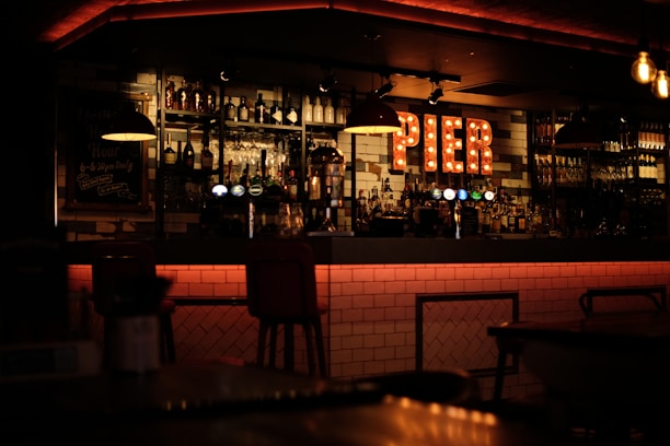 A warmly lit bar interior with shelves stocked with various bottles of liquor and glasses. A light-up sign spells 'PIER' and creates a cozy ambiance. The bar counter has a modern, tiled design, and there are several bar stools lined up in front. Hanging lights add to the subdued atmosphere.