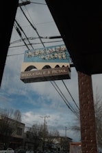 A vintage-style sign for 'Herkimer Coffee' hangs outside a brick building. Below the main sign, a smaller sign reads 'Wholesale & Retail.' The sign is positioned under a clear sky with some clouds, and power lines are visible running across the image. Bare trees and a street with buildings and cars can be seen in the background.