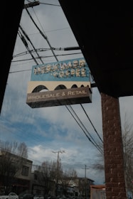A vintage-style sign for 'Herkimer Coffee' hangs outside a brick building. Below the main sign, a smaller sign reads 'Wholesale & Retail.' The sign is positioned under a clear sky with some clouds, and power lines are visible running across the image. Bare trees and a street with buildings and cars can be seen in the background.