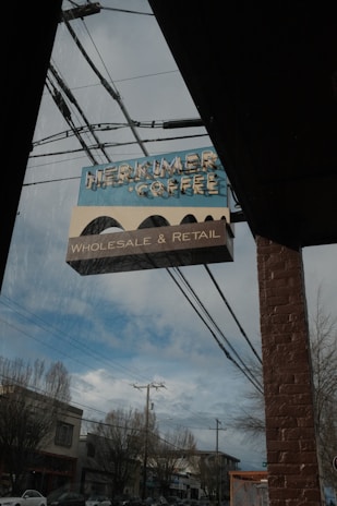 A vintage-style sign for 'Herkimer Coffee' hangs outside a brick building. Below the main sign, a smaller sign reads 'Wholesale & Retail.' The sign is positioned under a clear sky with some clouds, and power lines are visible running across the image. Bare trees and a street with buildings and cars can be seen in the background.