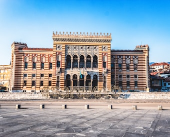 A large, ornate building with intricate architectural details, including a series of arches and patterned stonework. The building features a mix of colors, primarily shades of brown and beige, with decorative elements that add to its grandeur. In front of the building, there is a large sign spelling out 'Sarajevo' with a hashtag. The sky above is clear and blue.