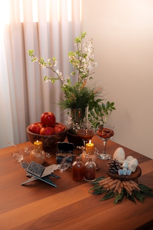 A wooden table is elegantly arranged with various items, including a bowl of red apples, lit candles, glass bottles, and a decorative vase with white and green foliage. There are also dried wheat stalks, a pine cone, and decorative eggs placed on a small wooden stand. The setting is warmly lit, creating a cozy atmosphere.