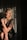 A person with blonde hair is sitting on a stool, wearing a vintage black dress adorned with gold accents. They have a headband with decorative elements and are holding a long beaded necklace. The lighting is dramatic, casting shadows on the light curtain backdrop.