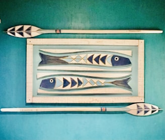 Wooden wall art features two stylized fish with geometric patterns, framed by two oars placed horizontally above and below on a teal background.