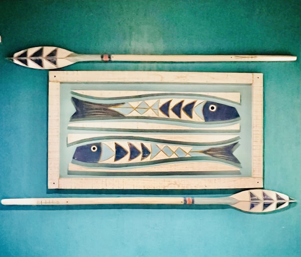 Wooden wall art features two stylized fish with geometric patterns, framed by two oars placed horizontally above and below on a teal background.