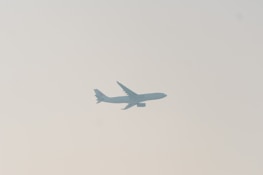 A commercial airplane is silhouetted against a pale sky, with its wings and tail clearly visible. The image conveys a sense of movement and travel.
