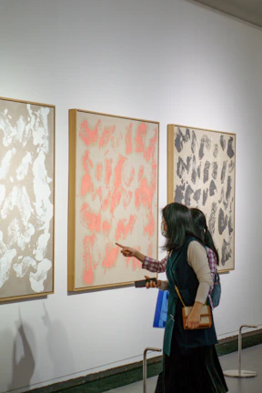 Two friends discussing a striking mixed-media queer art piece in the airy, white-walled gallery.
