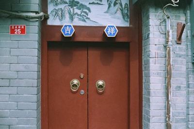 A red door with two brass handles is set within a brick wall. Above the door, two blue hexagonal plaques with white Chinese characters are displayed. There is a traditional Chinese painting above the door depicting a landscape. The brick wall appears gray, with some wear and tear visible. A red sign with white text and the number 25 is on the wall beside the door. Electrical conduits run along the side of the wall.