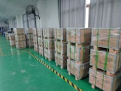Numerous wooden crates are stacked and wrapped with plastic on a factory floor. The boxes have green straps and are labeled as containing lithium-ion batteries. The floor is green, and the room appears to be spacious and well-lit, with large windows and ceiling fans.