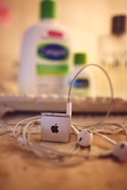 A set of white earphones is connected to a small silver device with an apple logo, resting on a textured surface. In the blurred background, there are bottles of lotion or cosmetic products with green caps and labels.