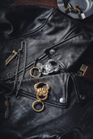A black leather jacket is prominently displayed with silver buttons and zippers. A gold and silver keychain with crab designs are placed on the jacket. There are also a feather pendant and a cross-like pendant on a chain, along with an ashtray containing a few bullets in the background.