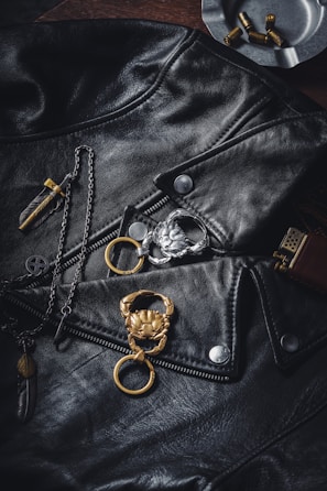 A black leather jacket is prominently displayed with silver buttons and zippers. A gold and silver keychain with crab designs are placed on the jacket. There are also a feather pendant and a cross-like pendant on a chain, along with an ashtray containing a few bullets in the background.