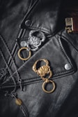 A black leather jacket is adorned with two crab-shaped keychains, one silver and one gold. There is also a chain necklace with a pendant in the shape of a feather. On the right, the top part of a brass-colored lighter is visible.