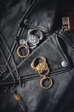 A black leather jacket is adorned with two crab-shaped keychains, one silver and one gold. There is also a chain necklace with a pendant in the shape of a feather. On the right, the top part of a brass-colored lighter is visible.