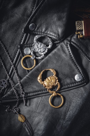 A black leather jacket is adorned with two crab-shaped keychains, one silver and one gold. There is also a chain necklace with a pendant in the shape of a feather. On the right, the top part of a brass-colored lighter is visible.
