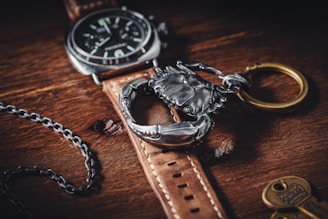 A leather watch strap with a silver watch face rests on a wooden surface. Next to the watch, a metallic crab-shaped keychain with a brass ring is prominently displayed. A dark metallic chain sits next to the watch.